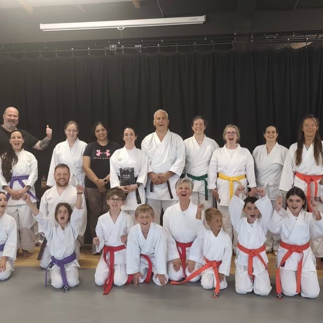 A mixed-age karate class poses in white uniforms in front of a black curtain.