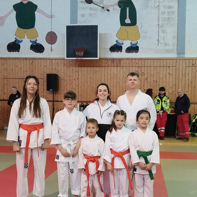 Adults and children in white karate uniforms pose together in a sports hall.