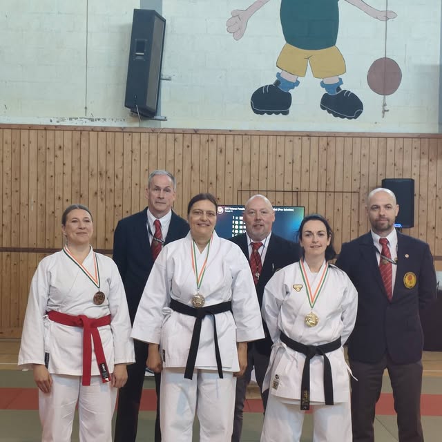 Three medal-wearing karate practitioners pose with officials in a sports hall.