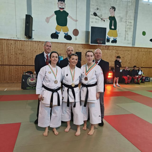 A group of medal-wearing karate practitioners and officials pose on red and green mats.