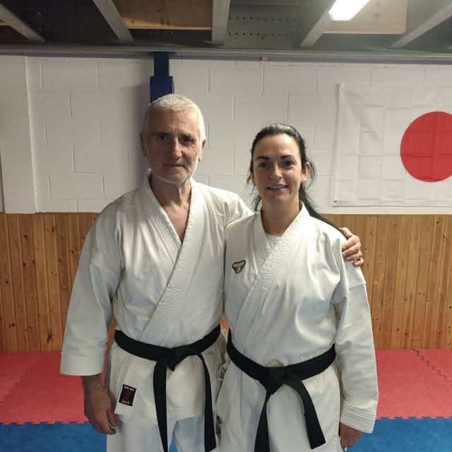 Two adult karate practitioners in white uniforms pose together on blue and red training mats.