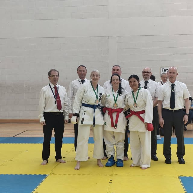 Medal-wearing karate practitioners and officials pose together on yellow and blue tournament mats.