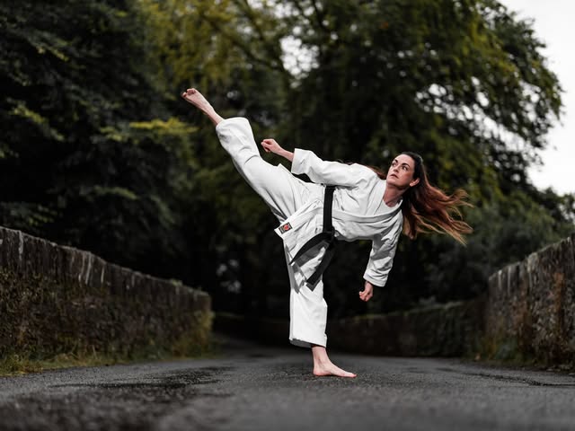 An adult karate practitioner performs a high side kick on a tree-lined road.